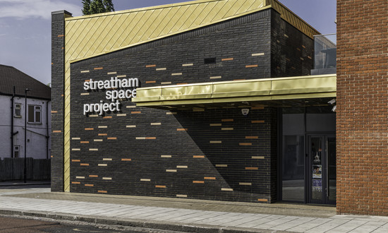 The exterior of Streatham Space Project, with black and red bricks and a yellow awning with Streatham Space Project signage lettering in white.